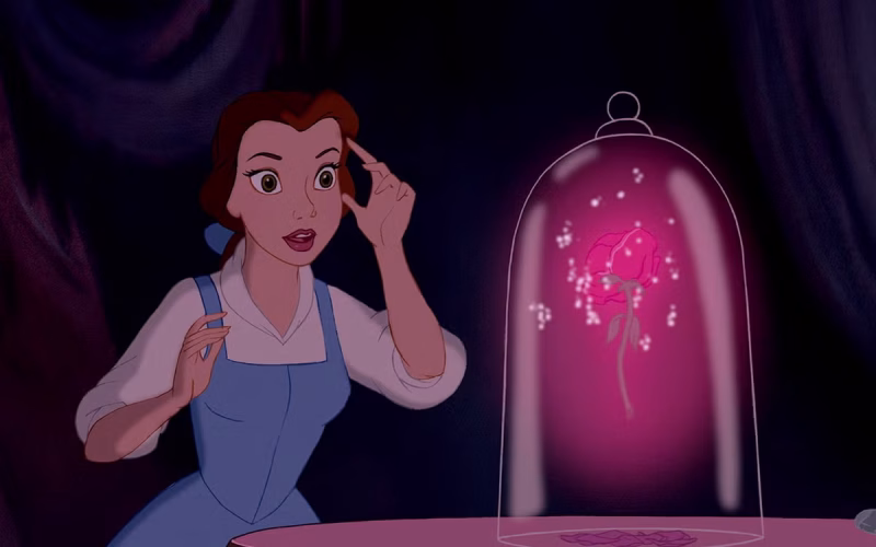 The character Belle from Disney's Beauty and the Beast looks with wonder at the glowing Enchanted Rose under a glass cloche.